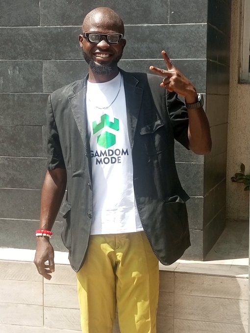 A smiling man wearing glasses, a black blazer, and yellow pants stands against a tiled wall, wearing a white t-shirt that reads "GAMDOM MODE" with a green logo, and flashing a peace sign.
