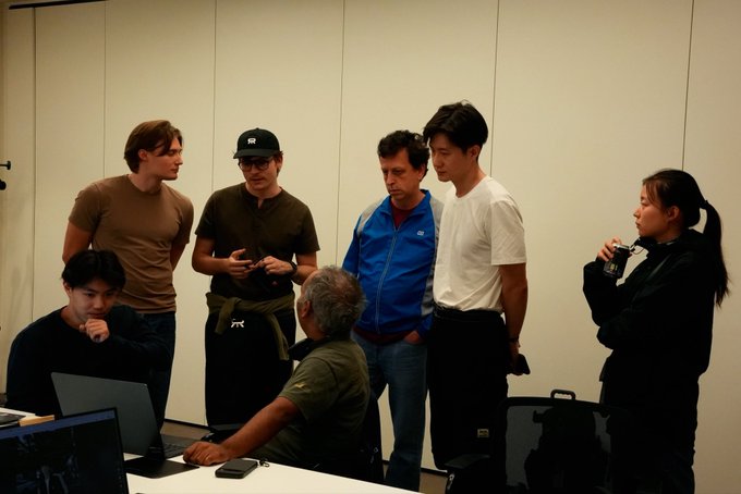 A group of hackers gathers around a laptop at the World Models London Hackathon, collaborating and reviewing projects together. One participant's cap features the "RW" logo of the sponsor @reactorworld, reflecting the community-driven spirit of the event.