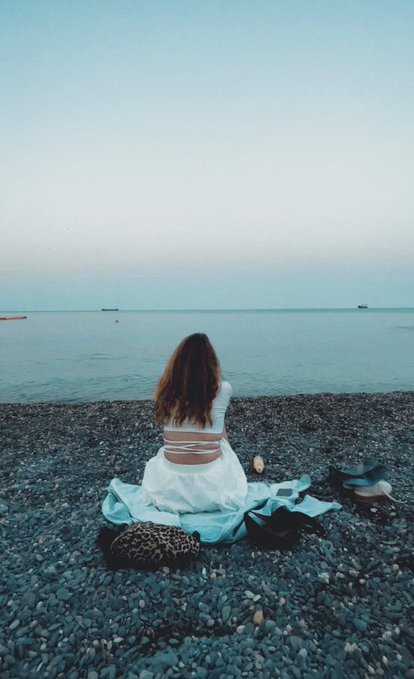 A woman in a white outfit sits on a rocky pebble beach facing a calm sea at dusk, with a turquoise blanket and bag beside her.