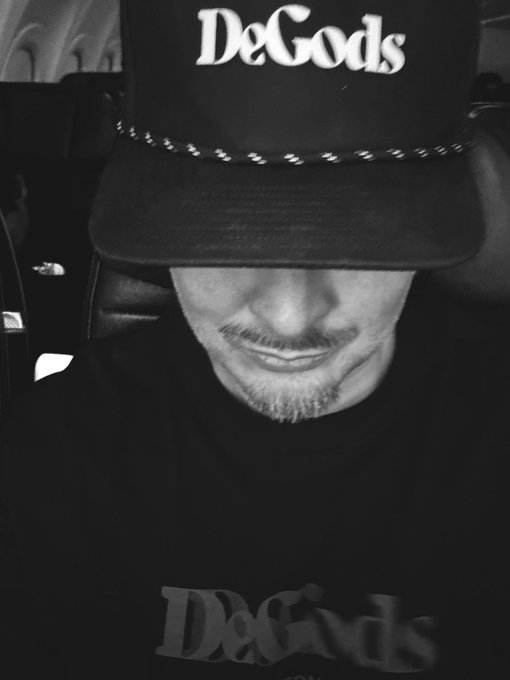 A black-and-white selfie of a man wearing a black baseball cap and a matching t-shirt, both bearing the text 'DeGods'. Only the lower half of his face is visible beneath the cap's brim.