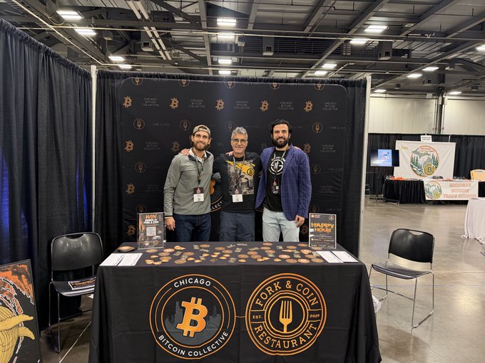 Three men pose behind a table with 'Chicago Bitcoin Collective' and 'Fork & Coin Restaurant' logos at a Bitcoin event, with books 'The Blocksize War' and 'Happy Bitcoin' displayed.