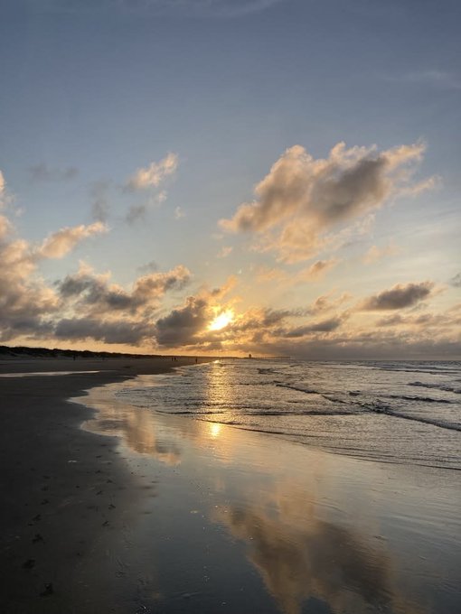 A serene beach at sunset features wet sand reflecting the sun, gentle waves rolling onto the shore, and scattered clouds in a blue sky.