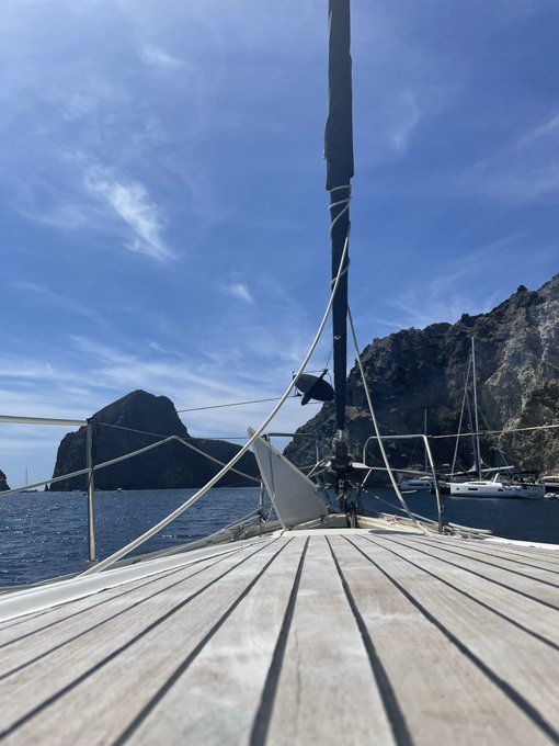 A view from the deck of a sailboat looking out at the ocean with rocky cliffs and other boats visible under a blue sky.