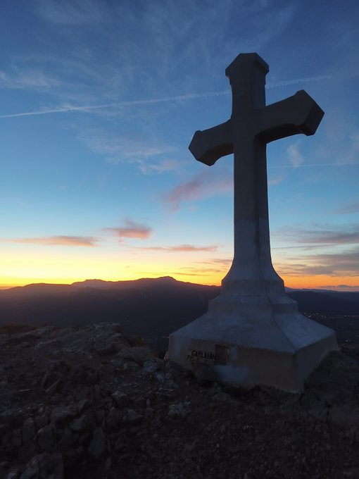 A large stone Christian cross silhouette stands atop a rocky mountain at sunset, with a plaque reading 'CAPILLA' visible at its base.