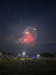 Red fireworks explode in the night sky above a grassy field where people are gathered to watch the display.
