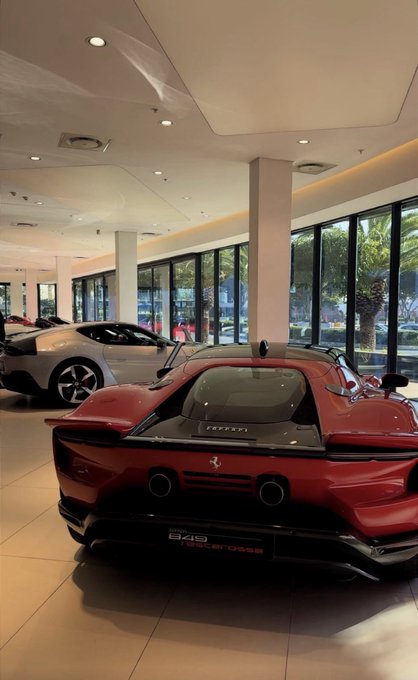 A red Ferrari sports car is displayed in a showroom, visible through a window with "GM" lettering and text reading "IT'S ONLY THE BEGINNING," "SALES HOURS," and "SERVICE HOURS." The luxury vehicle complements the post's message of manifesting big wins and success.