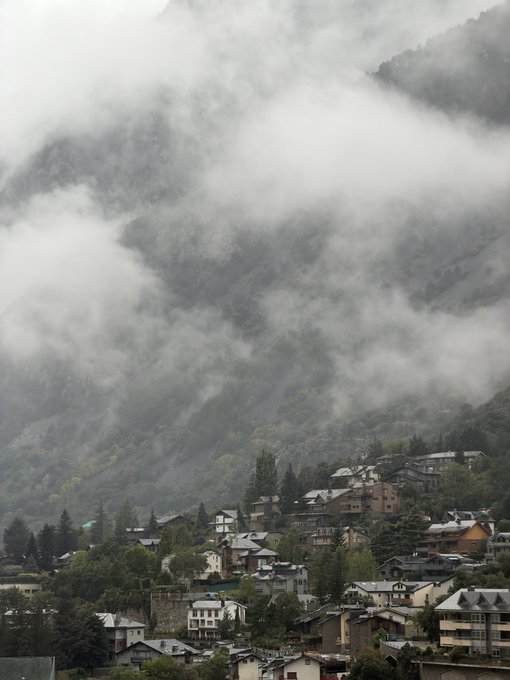 A mountain town is nestled at the base of steep, forested slopes shrouded in thick mist and low-hanging clouds. The moody, atmospheric scene evokes a peaceful, "good morning" vibe as the community begins its day under the dramatic, foggy mountain backdrop.