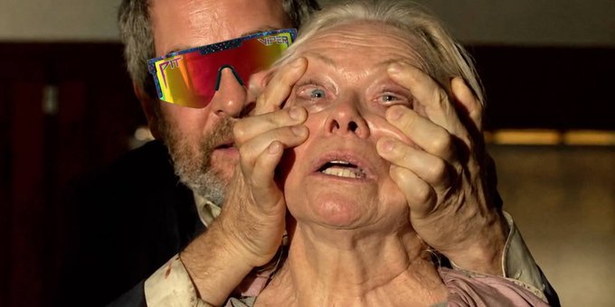 A man in colorful reflective sports sunglasses covering an elderly woman's eyes with his hands in a dimly lit setting.
