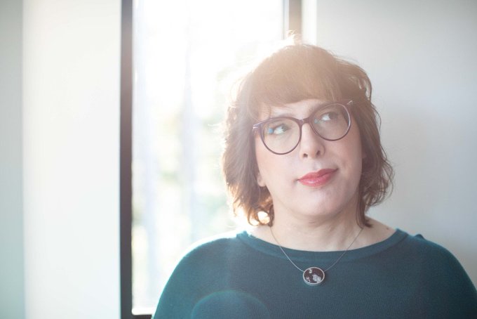 A close-up portrait of a woman with short reddish-brown hair, wearing glasses, a green sweater, and a Texas-shaped pendant necklace, lit by a bright window.
