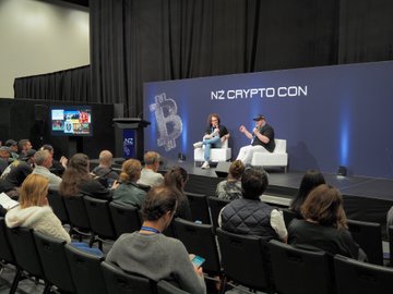 The image shows a large group of people sitting in chairs, watching two men stand behind them at a podium. One man is wearing a black shirt while the other has a white hat. The audience appears engaged as they listen to the speakers discussing real dogecoin utility and its potential impact on the cryptocurrency market.