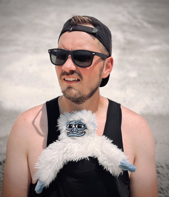 A man wearing sunglasses, a backwards black cap, and a black tank top holds a white fluffy "$LOFI" plush toy tucked into his shirt, visually representing the caption's claim that the token now "lives" in his shirt while crediting it for his journey into crypto, content creation, and web3.