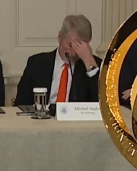 A man in a dark suit and red tie sits at a conference table with his hand covering his face in apparent frustration, with a visible nameplate reading "Michael Saylor." The image reflects the post's commentary on Saylor's poor market timing, capturing the look of dismay fitting the joke about buying high and selling low.
