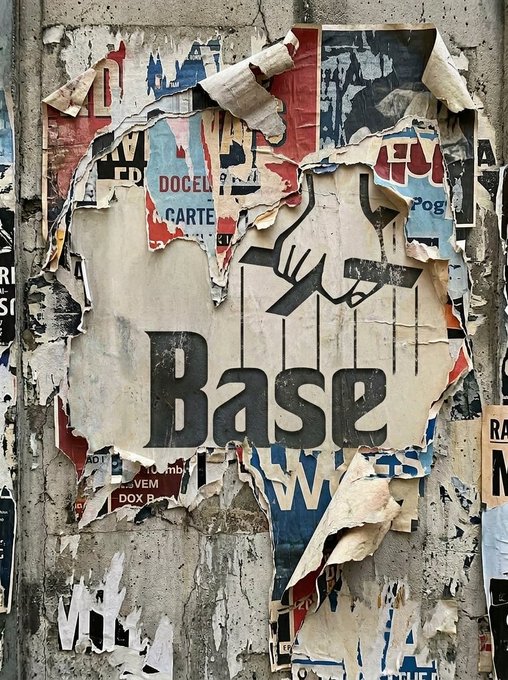 A weathered, peeling poster on a concrete wall displays the word 'Base' in large black letters, surrounded by remnants of other torn posters showing partial words like 'DOCEL', 'CARTE', and 'OVEM'.