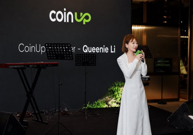 A woman in a long white dress speaking into a microphone at a CoinUp event, with the CoinUp logo and text 'CoinUp Speaker - Queenie Li' displayed on the screen behind her alongside a keyboard setup.
