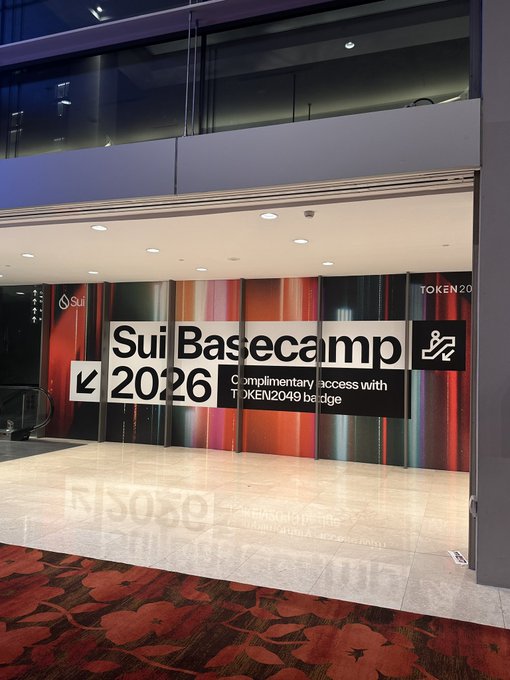 A glass entrance to "Sui Basecamp 2026" with a TOKEN2049 badge requirement sign, featuring a colorful striped backdrop and a directional arrow, captured as the poster gears up to share alpha from the @SuiNetwork event.