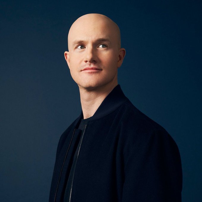 A professional portrait photograph of a bald man wearing a dark navy jacket, photographed against a solid dark blue background.