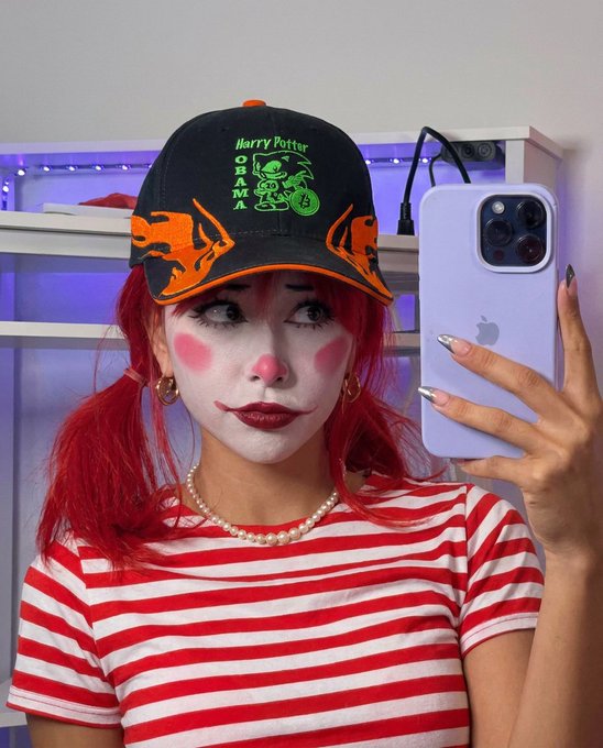 A red-haired woman taking a mirror selfie in clown makeup (white face, red cheeks/nose, drawn smile) wearing a red-and-white striped shirt and a black "Harry Potter" cap with orange bucking broncos.
