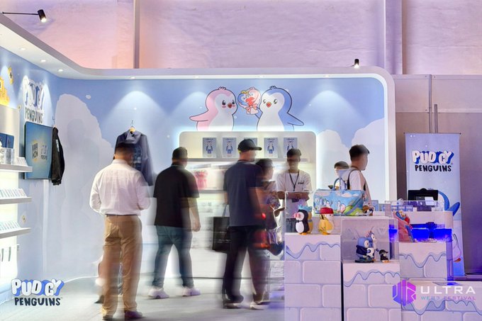 A Pudgy Penguins merchandise booth with people browsing products, branded with 'Pudgy Penguins' and 'ULTRA' logos.