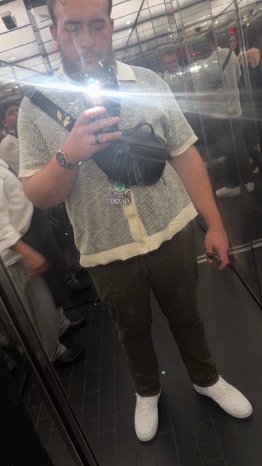 A mirror selfie taken in an elevator shows a man wearing a white short-sleeved knit shirt, dark pants, and white sneakers, holding a phone and wearing a black crossbody bag.