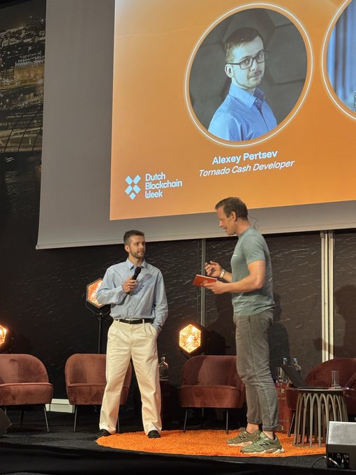 In the image, two men are standing next to each other on stage with a large screen behind them displaying an orange background featuring a man's face and the words "Dutch blockchain Turno Cash Developer" written above him. One of the men is holding a microphone while the other one has a book in his hand. The scene suggests that they might be participating in a panel discussion or interview, possibly related to the development of the Dutch blockchain technology for cash transactions.