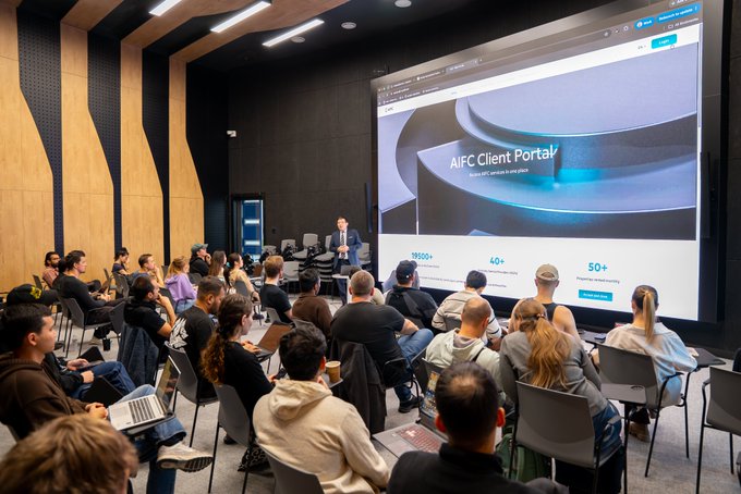 A presentation in a modern lecture hall where a speaker addresses an audience seated in rows, with a large screen displaying "AIFC Client Portal" and statistics like "13000+", "40+", and "50+".