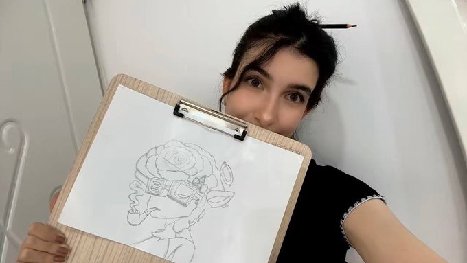 A woman with a pencil behind her ear holds up a clipboard displaying a pencil sketch of a character with VR goggles and a rose for hair, partially covering her face.