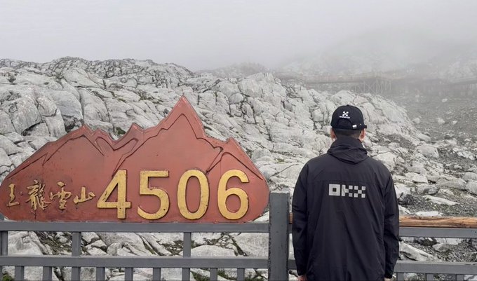 A man wearing a black jacket and cap stands before a sign reading '玉龍雪山 4506' (Jade Dragon Snow Mountain 4506m) with a rocky, foggy landscape in the background.