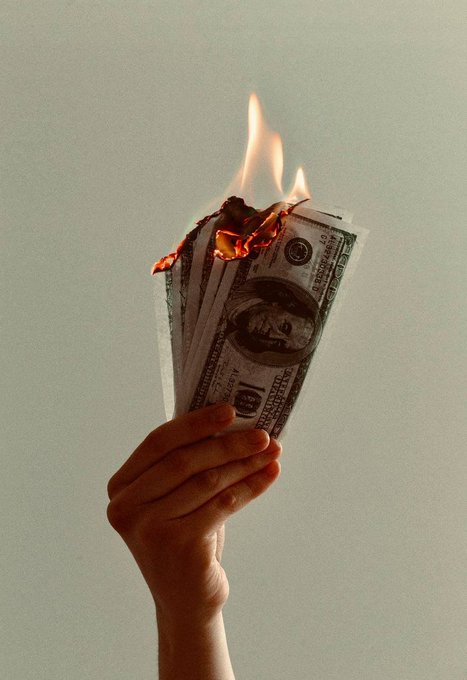 A hand holds a fan of U.S. one-hundred-dollar bills that are engulfed in flames against a pale background.
