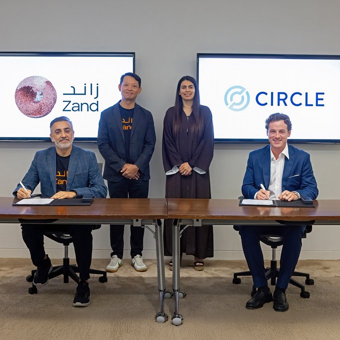 Four people pose for a photo at a signing ceremony, with two men seated at a table signing documents while a man and woman stand behind them; screens display logos for 'Zand' and 'CIRCLE'.