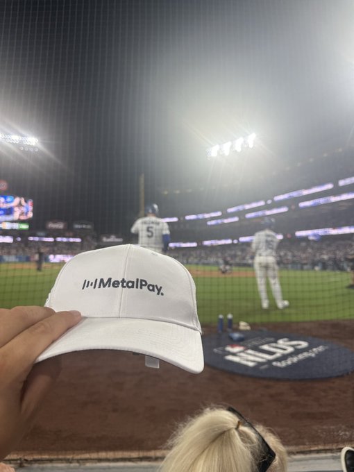 A photo taken at a baseball game where a hand is holding up a white 'MetalPay' branded cap in the foreground, with the game in progress in the background.
