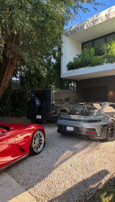 A collection of luxury vehicles parked on a gravel driveway outside a modern home, featuring a red Ferrari, a gray Porsche 911 GT3, and a black Mercedes-Benz G-Wagon.