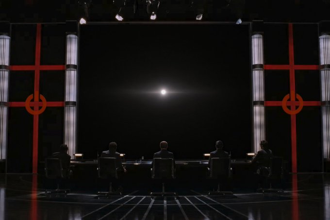 Five silhouetted figures sit at a long conference table in a dark, imposing chamber, gazing toward a single bright light source in the center of the black wall ahead, flanked by red geometric architectural elements.