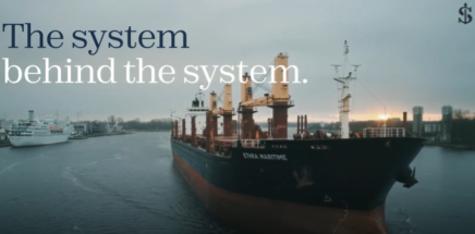 A large cargo ship docked in a harbor at sunset with the overlaid text 'The system behind the system.'