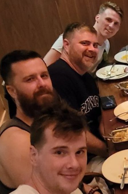 A selfie of four men smiling at the camera while seated at a restaurant table with plates of food and drinks visible.