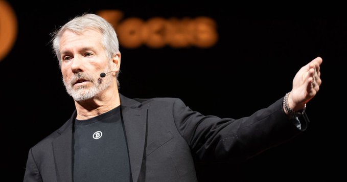 A photograph of a gray-haired man in a black blazer and dark shirt, wearing a headset microphone, gesturing with his hand while speaking on a stage with orange text visible in the background.
