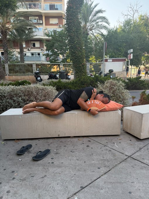 A man lies horizontally across a concrete bench in an urban plaza, fanning himself with an orange hand fan reading "IN A FAN" while wearing shorts and a t-shirt, with his flip-flops abandoned on the pavement below.

The image humorously captures the "bear market vibes" by depicting someone utterly exhausted and defeated, fanning themselves in the heat as if they have nothing left to give—mirroring the drained, languid feeling many investors experience during a prolonged crypto downturn.