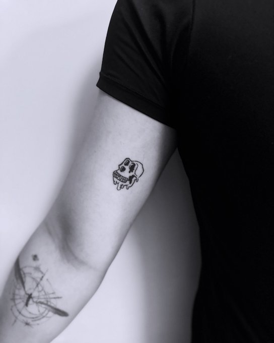 A black-and-white photograph showing a person's upper arm with a small tattoo of a pug dog, with another tattoo of intersecting lines visible on the forearm below.