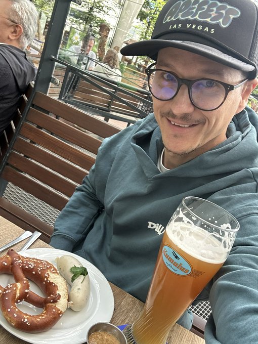 The image features a man sitting at an outdoor table with a plate of pretzels, a glass of beer, and a hat on his head. The man appears to be enjoying his meal while wearing a hat that says "Las Vegas". In the background, there are other people present in the scene, suggesting that this is a public space or gathering area.