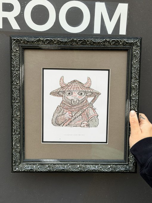 A framed artwork in an ornate dark frame displayed under a 'ROOM' sign, showing a detailed illustration of a horned creature wearing a traditional Asian conical hat and robe, with a hand visible holding the frame.