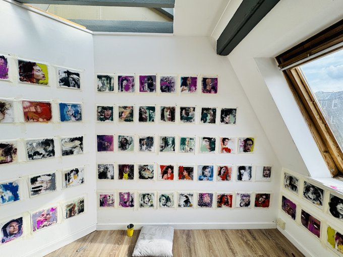 An art studio with white walls displaying numerous small portrait paintings hung in rows using masking tape, featuring colorful painted faces in various styles, with a skylight above and a floor cushion in the center.