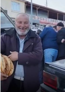A smiling, bearded man in a dark coat proudly holds up what appears to be a bundle of bread loaves, beaming broadly with joy as he stands near a car. The image humorously illustrates the poster's enthusiasm about scoring a great deal—likely comparing the "handing out boards at ridiculous prices" to receiving free bread, with the man's big grin symbolizing the anticipated satisfaction of closing the trade at the target price.