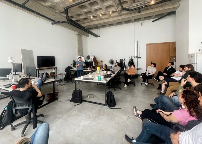 A group of people sit in a modern industrial-style office space with exposed concrete ceiling, listening to a presentation given by a standing man.