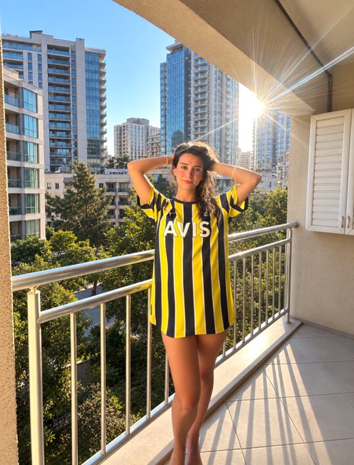 A woman stands on a balcony wearing an oversized yellow and black striped 'AVIS' jersey dress, posing with her hands in her hair against a city skyline.