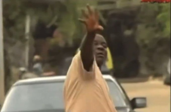 Blurry photo/video still of a man in an orange shirt gesturing with his raised hand on a street with cars in the background.