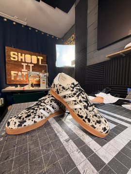 A pair of custom cow-print Adidas sneakers sits on a checkered surface in front of a neon "SHOOT IT" sign, displayed as the prize for the Honey Bee Lounge one-year anniversary giveaway. The sneakers feature a distinctive black-and-white cowhide pattern with gum soles, matching the giveaway theme of celebrating the lounge's milestone.