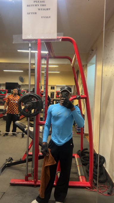 A man wearing a light blue long-sleeve shirt and black pants takes a mirror selfie at a gym while standing inside a red power rack beneath a sign reading 'PLEASE RETUN THE WEIGHT AFTER USAGE'.