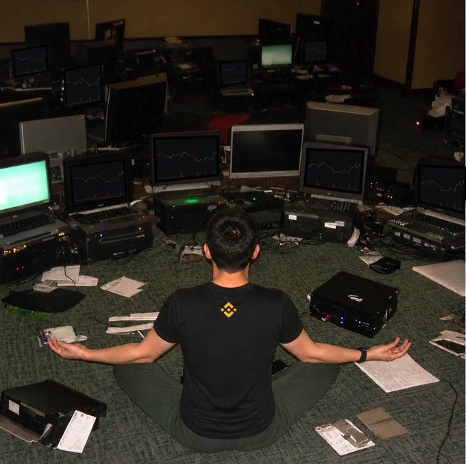 A person wearing a Binance-branded t-shirt meditates in a cross-legged lotus position, surrounded by multiple monitors displaying cryptocurrency trading charts in what appears to be a trading office or workspace, perfectly embodying the "Pause and breathe" message amid the chaos of trading.