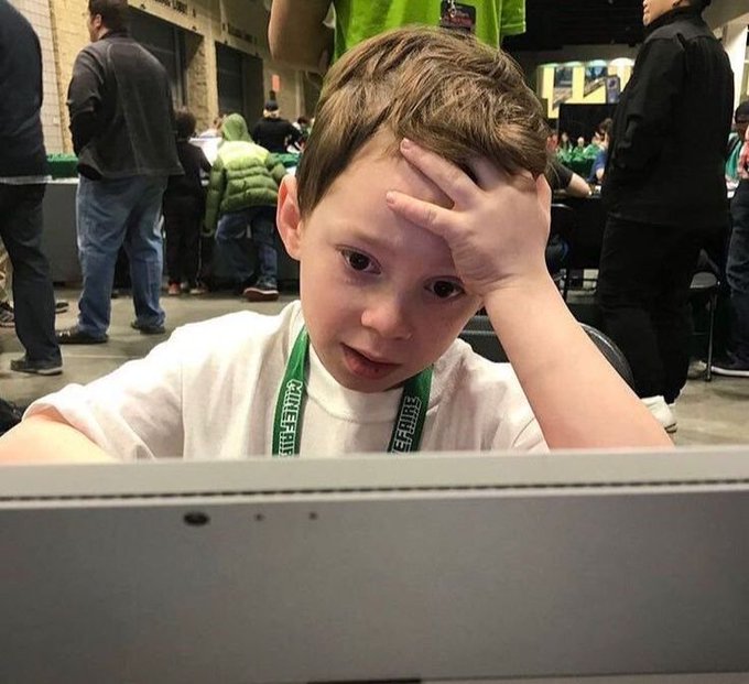 A young boy with a confused expression sits in front of a laptop, resting his hand on his forehead, with a green lanyard around his neck at what appears to be a gaming tournament.