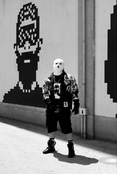 Black and white street photography image of a person wearing a white ski mask, patterned jacket, and black shorts standing in front of a wall featuring large pixel-art graffiti portraits.