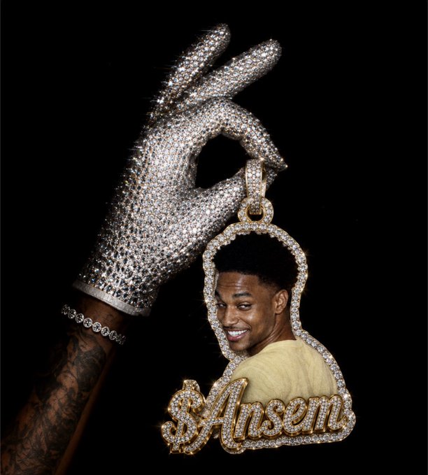 A rhinestone-encrusted hand holding a pendant featuring a portrait of a smiling man with the text '$Ansem' in gold lettering.